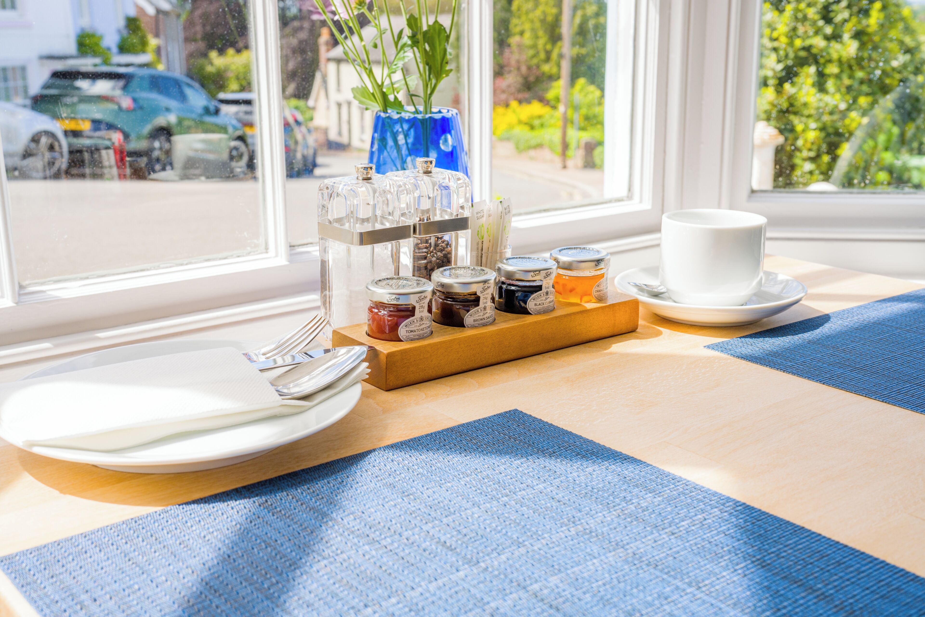 A breakfast setup with plates, cutlery, a tray of condiments, and a cup on a table by a window with a view of parked cars and greenery.