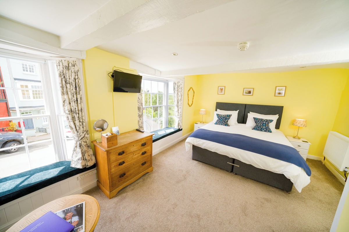 A bedroom with a bed, dresser, bedside table, lamp, window with curtains, and a view of the street.