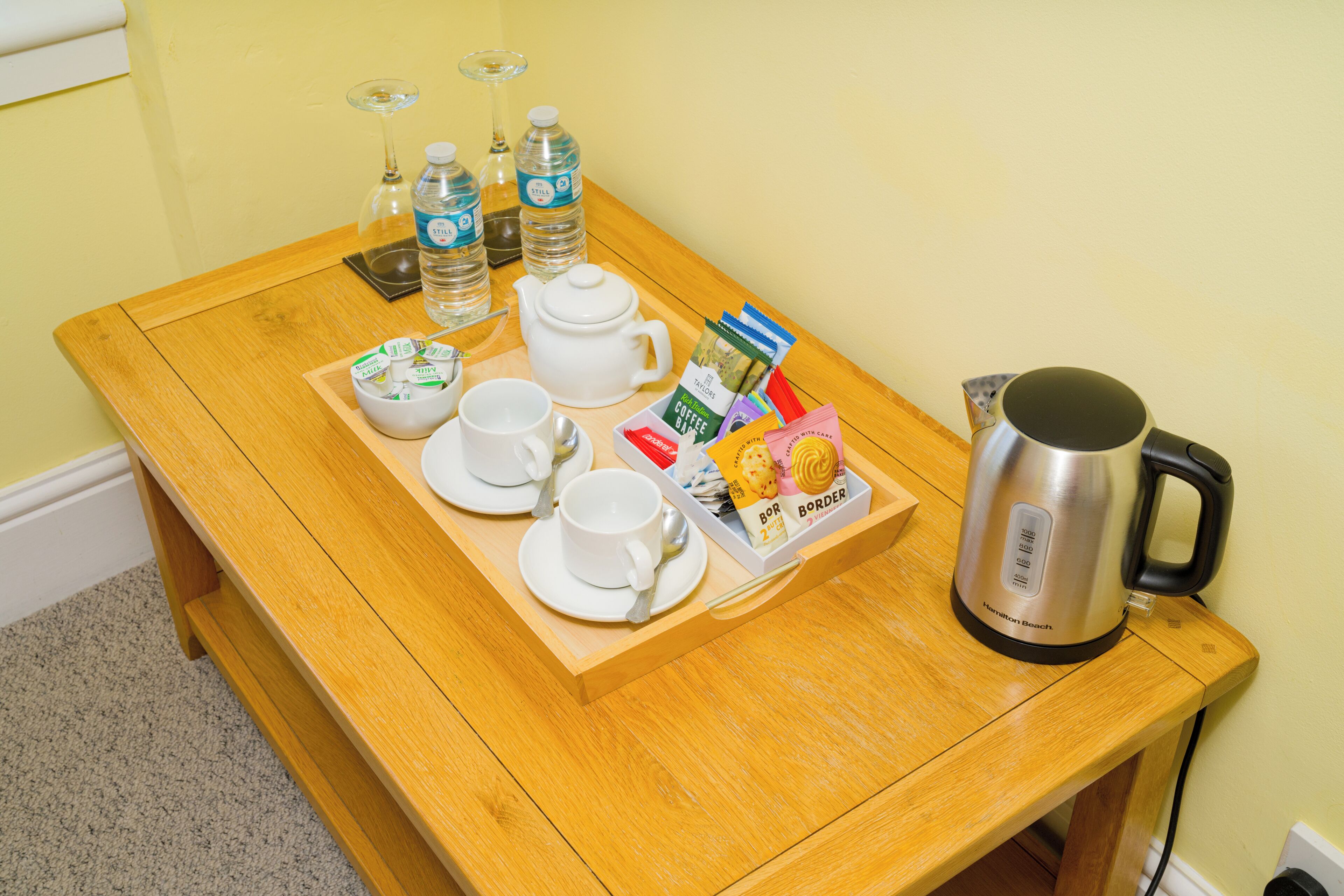 Una mesa de madera con una bandeja que contiene una tetera, tazas, un vaso, botellas de agua y una bandeja para café.