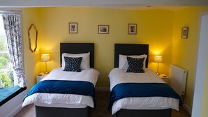 A double room with two beds, each with a headboard, white bedding, and blue throw pillows. There are bedside lamps, a window with curtains, and a radiator on the wall.