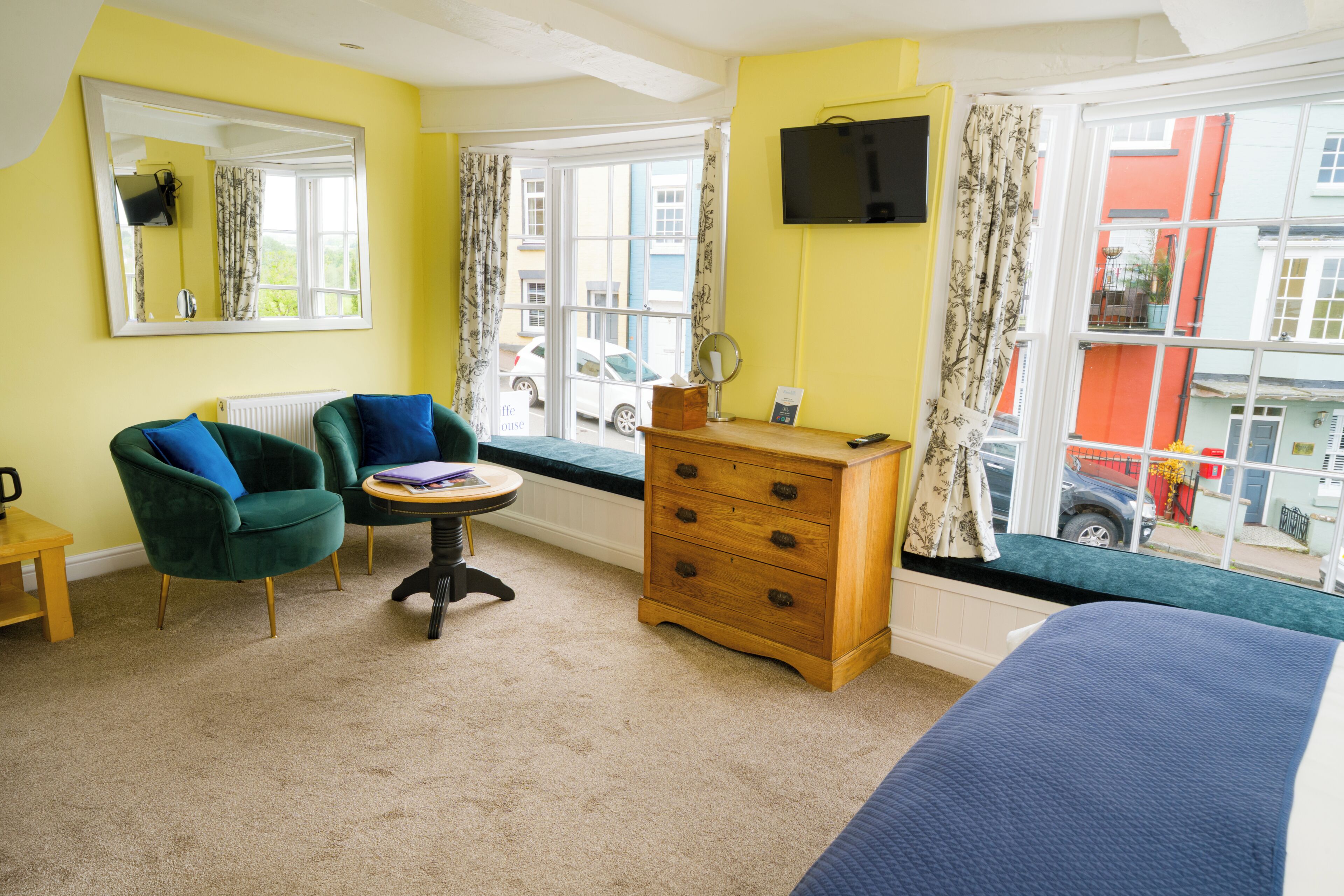 A room with a bed, a dresser, a chair, a small table, a mirror, and a window with a view of the street.