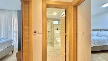 Una habitación de hotel moderna con una puerta de madera que da a un baño, una cama con ropa de cama blanca y una ventana con cortinas traslúcidas.