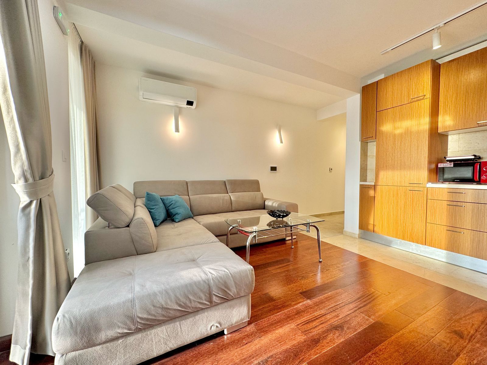 A modern living room with a beige sectional sofa, a glass coffee table, and a wooden floor. There is a kitchen area with wooden cabinets and a microwave on the counter.