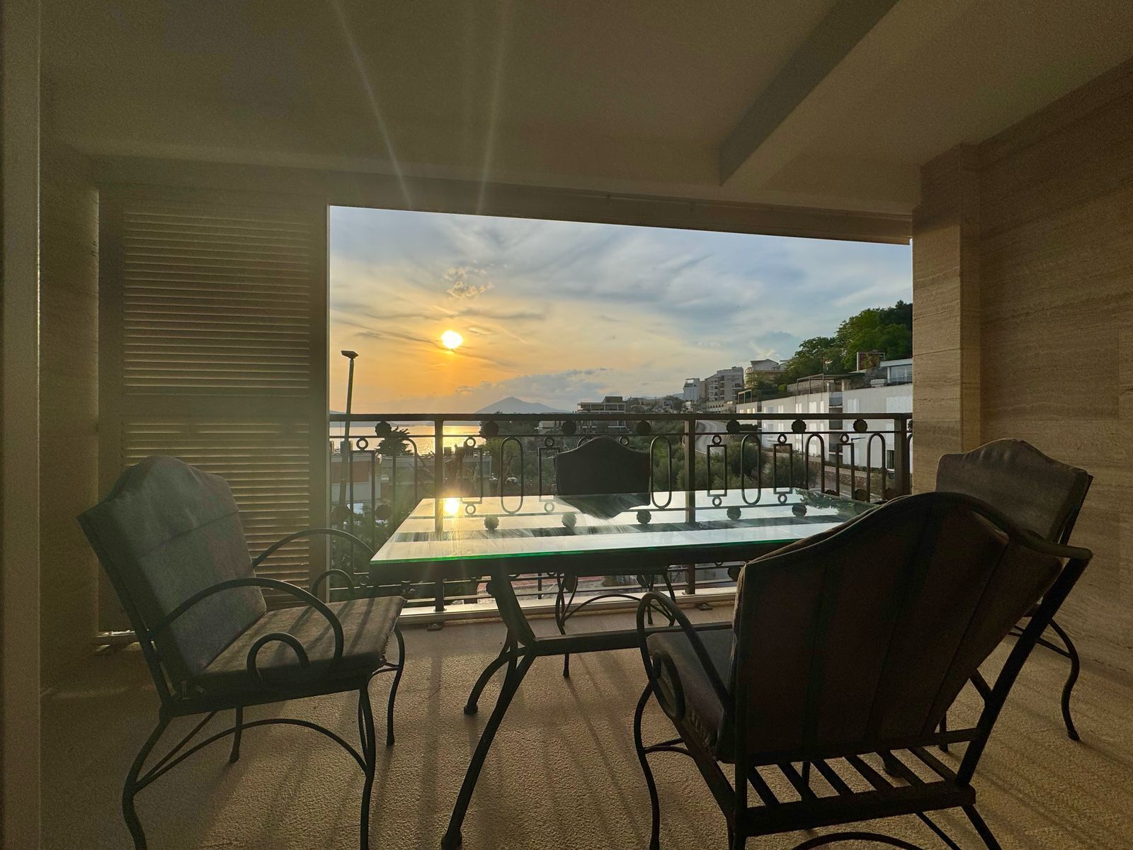 Un balcón con una mesa y sillas de vidrio, con vistas a un atardecer sobre un paisaje urbano.
