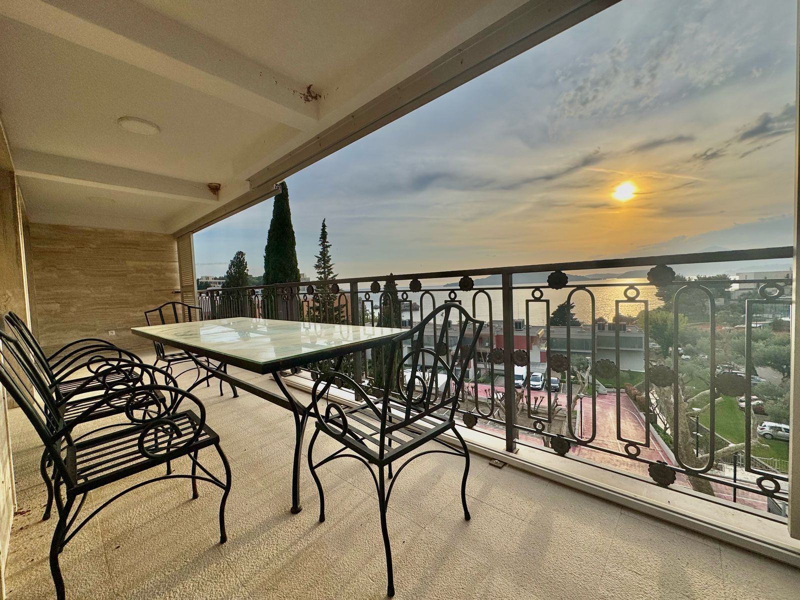 A balcony with a table and chairs, overlooking a cityscape and a body of water during sunset.