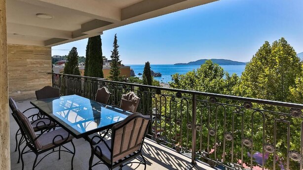 A balcony with a glass table and chairs, overlooking a scenic coastal view with trees and mountains.