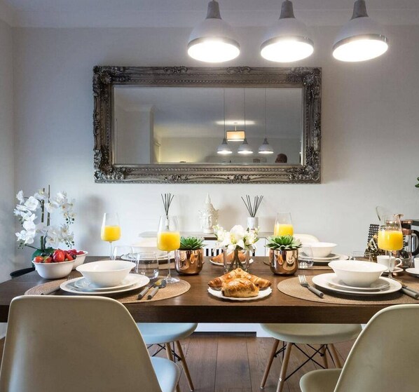 A dining area with a large mirror, three pendant lights, and a breakfast setting with croissants, fruit, and orange juice. - Westbourne Terrace Apartment in London Paddington (London)