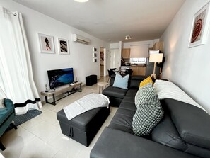 Ein modernes Wohnzimmer mit einem großen, schwarzen Sofa, einem Flachbildfernseher, einem kleinen Couchtisch und einem Essbereich im Hintergrund.