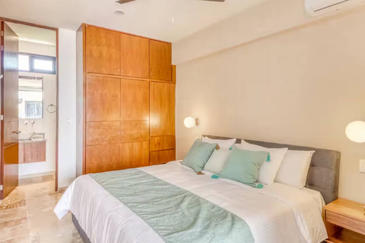 A bedroom with a bed, wooden wardrobe, bedside table, and a bathroom visible through an open door.