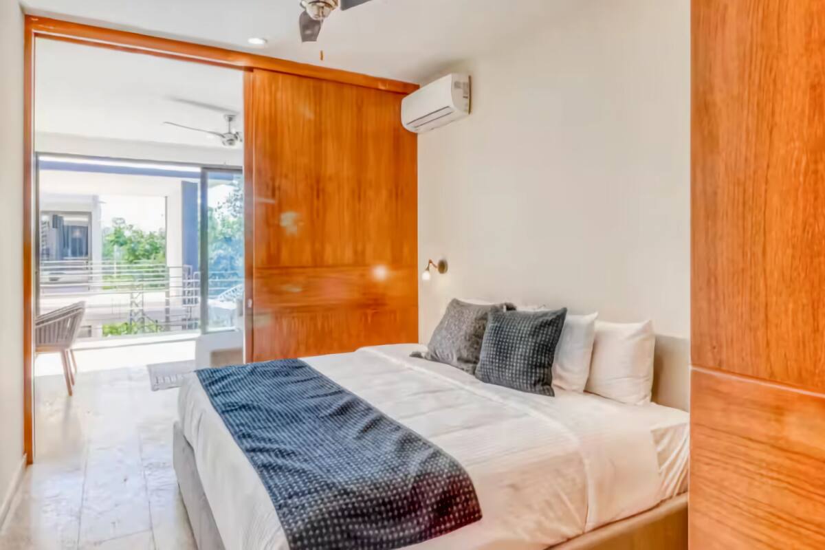 A bedroom with a bed, a wooden wardrobe, and a balcony with a view of greenery.