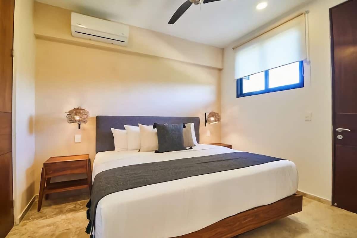 A bedroom with a bed, an air conditioning unit, a ceiling fan, a nightstand, and a window with blinds.
