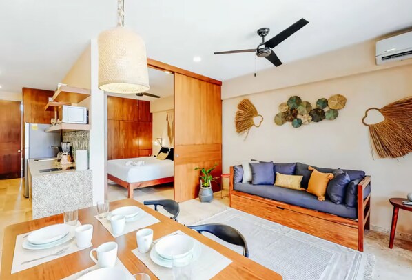 Una sala de estar moderna con una mesa de comedor de madera dispuesta para cuatro, un sofá, un ventilador de techo y un área de cocina visible al fondo.