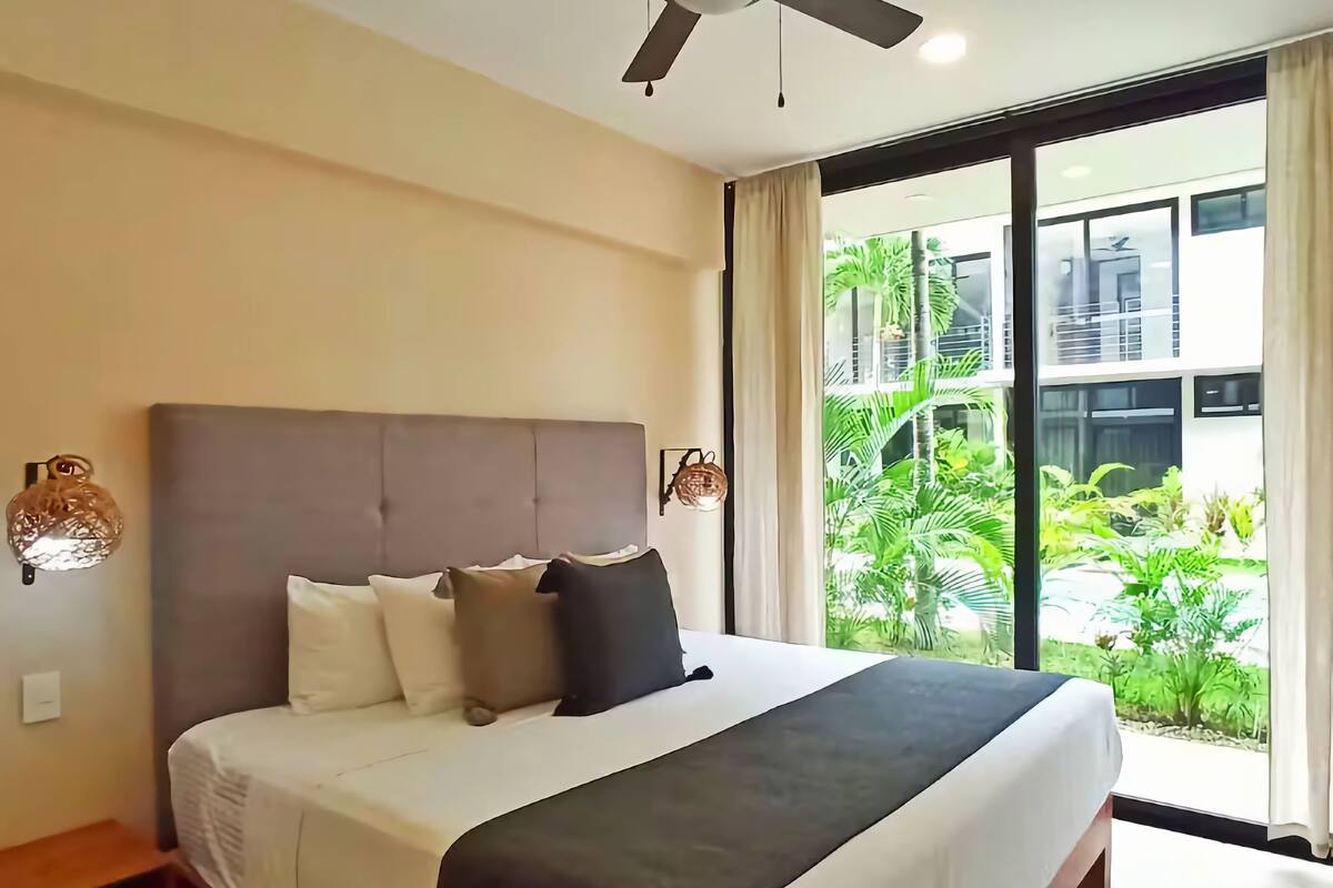A bedroom with a large bed, a ceiling fan, and a view of greenery outside.