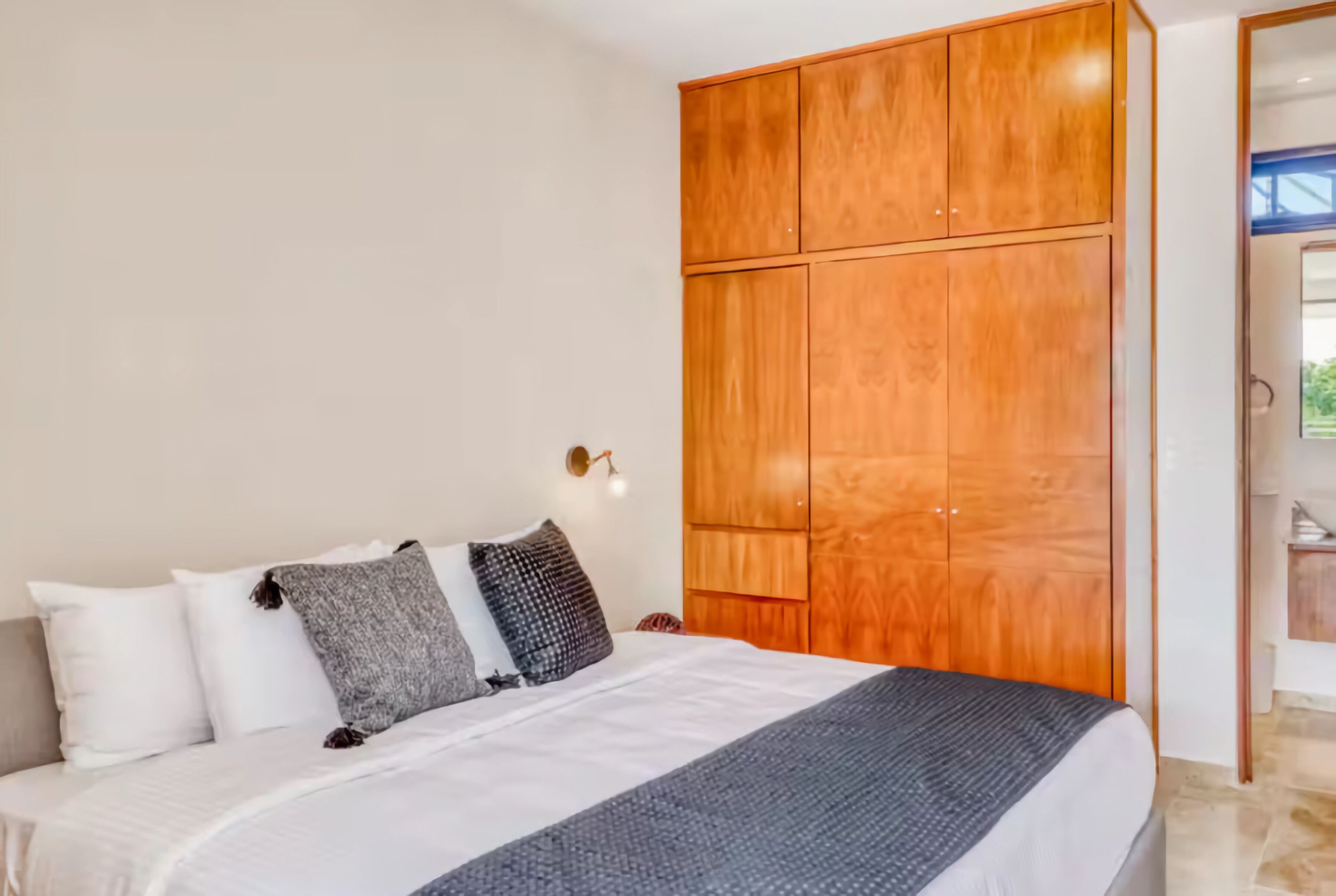 A bedroom with a bed, white and gray pillows, a wooden wardrobe, and a wall-mounted light fixture.