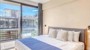 A modern hotel room with a large bed, a balcony with a view of buildings, and a large window.