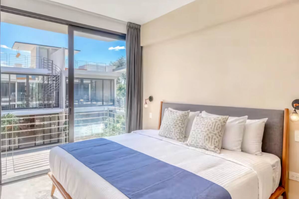 A modern hotel room with a large bed, a balcony with a view of buildings, and a large window.