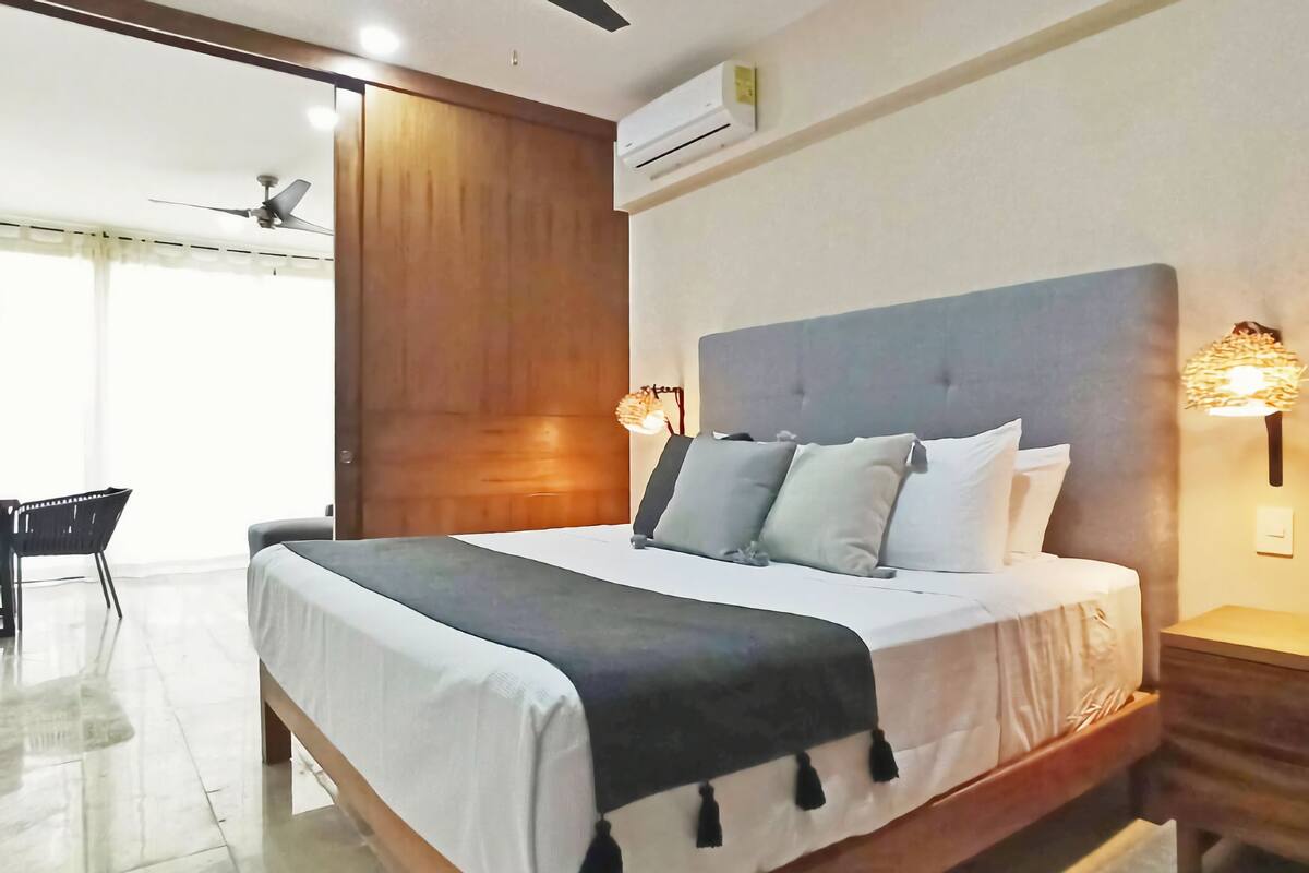 A bedroom with a large bed, a ceiling fan, an air conditioner, and a desk with a chair.