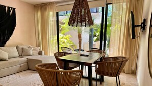 A modern living room with a sofa, a dining table with chairs, a large window with curtains, and a hanging light fixture.