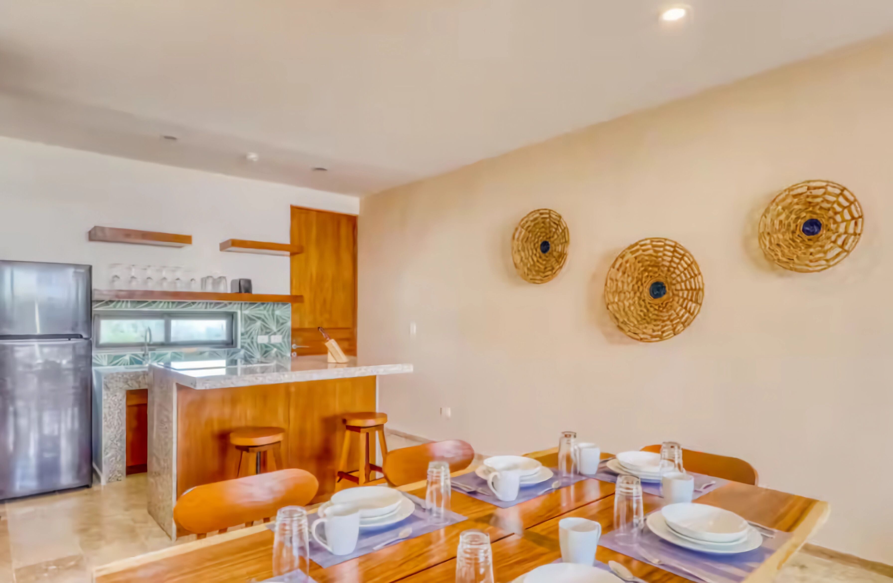 A modern kitchen with a central island, wooden bar stools, and a dining area set with white plates, cups, and glasses.