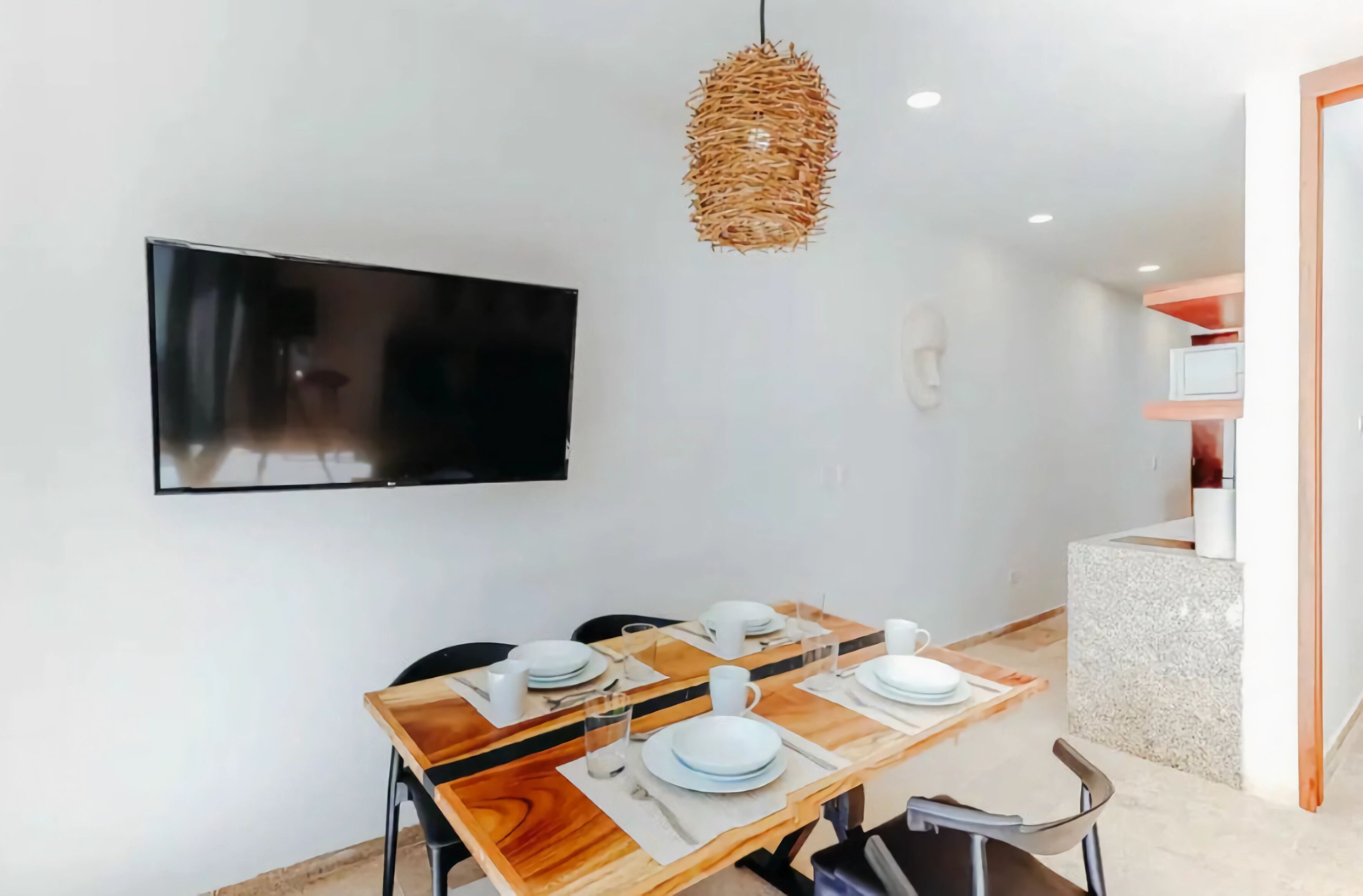 A dining area with a wooden table set for four, a flat-screen TV mounted on the wall, and a woven pendant light.