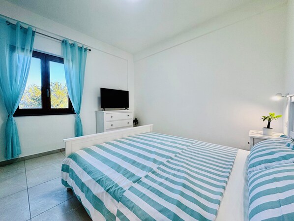 A bedroom with a bed, a TV on a dresser, a window with curtains, and a bedside plant.