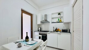 A compact kitchen with white cabinetry, a gas stove, a microwave, and a sink. There is a dining table set with plates, cups, and a bottle of wine. - YourHome - Relais Sant'Anna (Vico Equense)