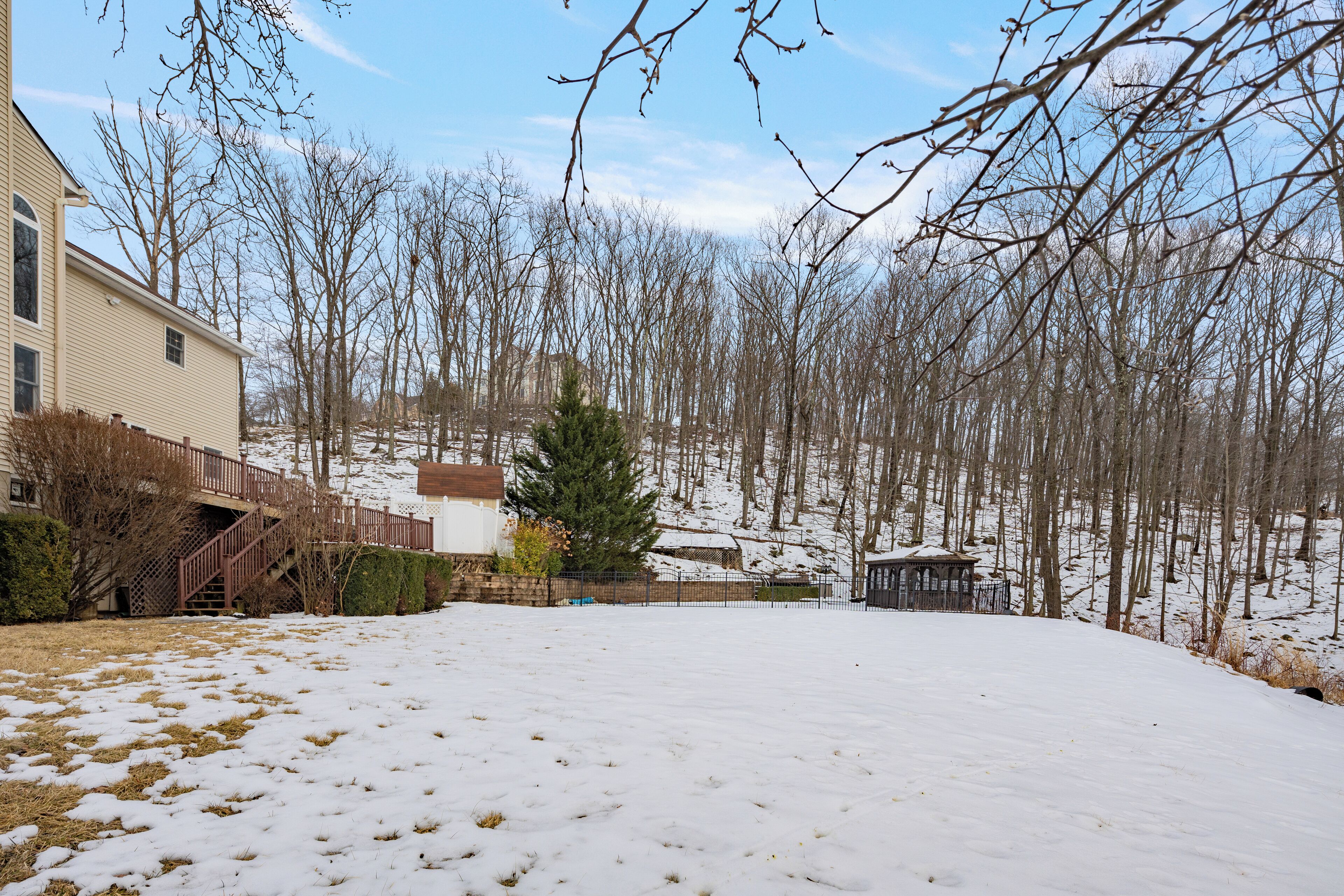 A snow-covered yard with a wooden deck, a small gazebo, and leafless trees.