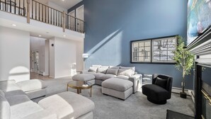 Una sala de estar moderna con un sofá modular, un sillón negro y una mesa de centro. Se ve una escalera que conduce a un nivel superior, y un cuadro grande en la pared.