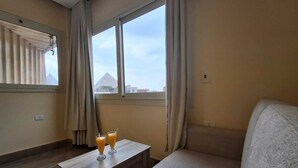 A room with a sofa, a small table with two glasses of orange juice, and a window with a view of a pyramid. - Hana Giza Pyramids Inn (Giza)