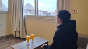 A person sitting at a table with three glasses of orange juice, a window with curtains, and a view of buildings and a mountain in the background. - Hana Giza Pyramids Inn (Giza)
