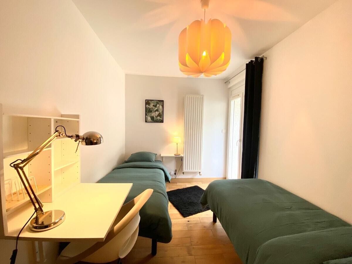 Une chambre avec deux lits simples, un bureau avec une lampe, un radiateur et une fenêtre avec des rideaux.