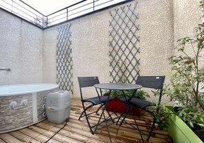 Un petit patio extérieur avec une table ronde et deux chaises, un jacuzzi et un bac à fleurs vert contenant des plantes.