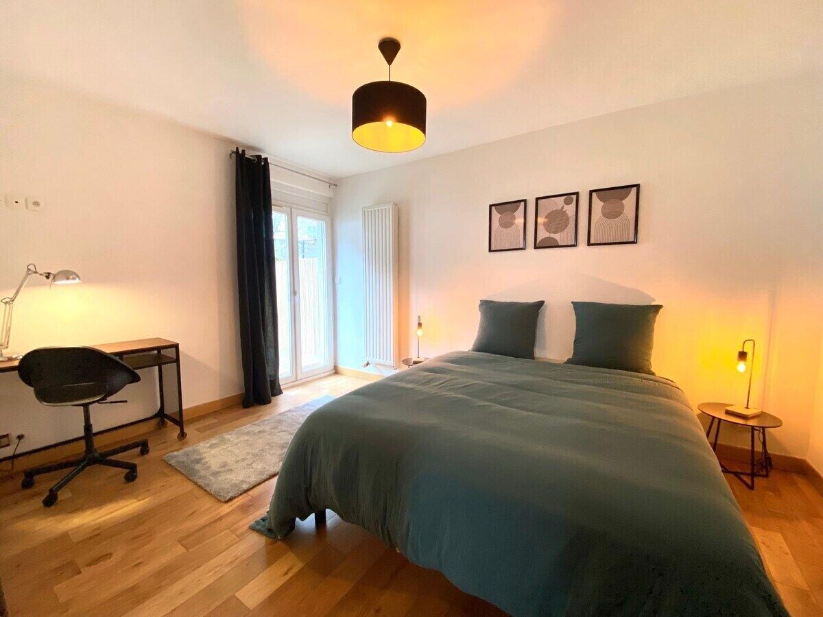 A bedroom with a bed, a desk with a chair, a standing lamp, and a window with curtains.