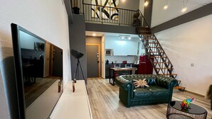 A modern living room with a flat-screen TV, a leather sofa, a dining table, and a staircase leading to an upper level.