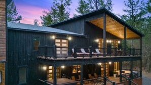 A two-story log cabin with a balcony, outdoor seating, and a stone foundation. - Kid's Play Room | Game Room | Hot Tub | Sleeps 22 (Broken Bow)