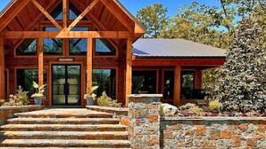 A wooden lodge with a steeply pitched roof, large windows, and a stone staircase leading to the entrance. - Kid's Play Room | Game Room | Hot Tub | Sleeps 22 (Broken Bow)
