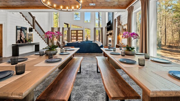 A spacious dining area with wooden tables and benches, large windows offering a view of the forest, and a chandelier hanging from the ceiling. - Kid's Play Room | Game Room | Hot Tub | Sleeps 22 (Broken Bow)
