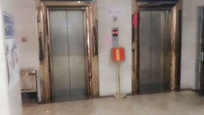 Un pasillo con dos ascensores, un botón de emergencia rojo y una pequeña mesa con una silla.