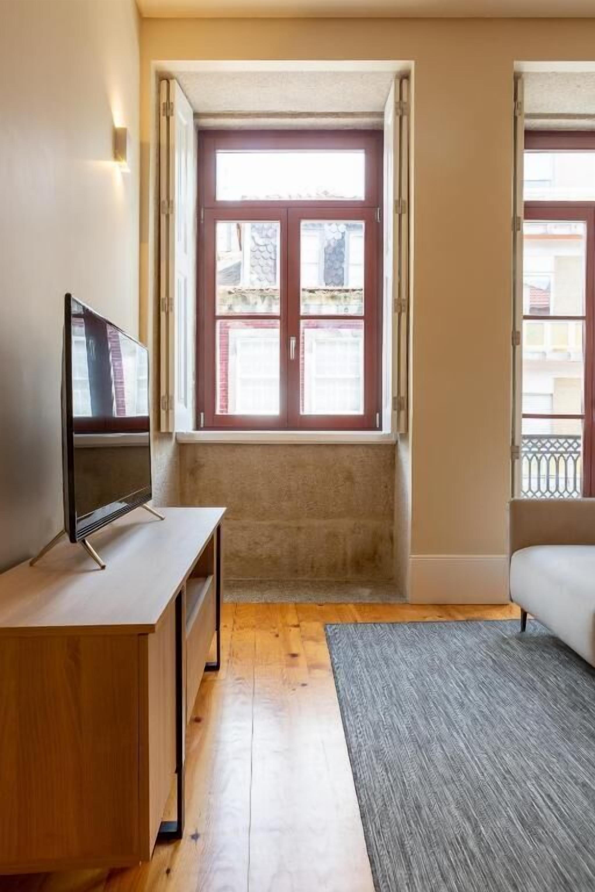 Een kamer met een houten bureau, een televisie, een grijs vloerkleed en een raam met uitzicht op gebouwen.