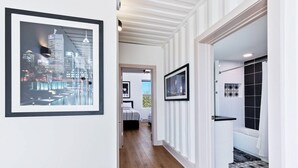 Un pasillo con arte urbano enmarcados, piso de madera y vista a un dormitorio con cama y ventana.