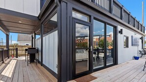Un edificio moderno con terraza de madera, grandes puertas correderas de vidrio y un área de estar al aire libre visible.