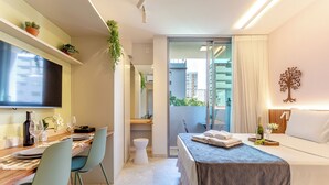 A modern hotel room with a dining area, a bed with a blue bedspread, a balcony with a view of buildings, and a wall-mounted tree decoration. - Moderno-Pinheiros -AC - Brazilian Corner (São Paulo)