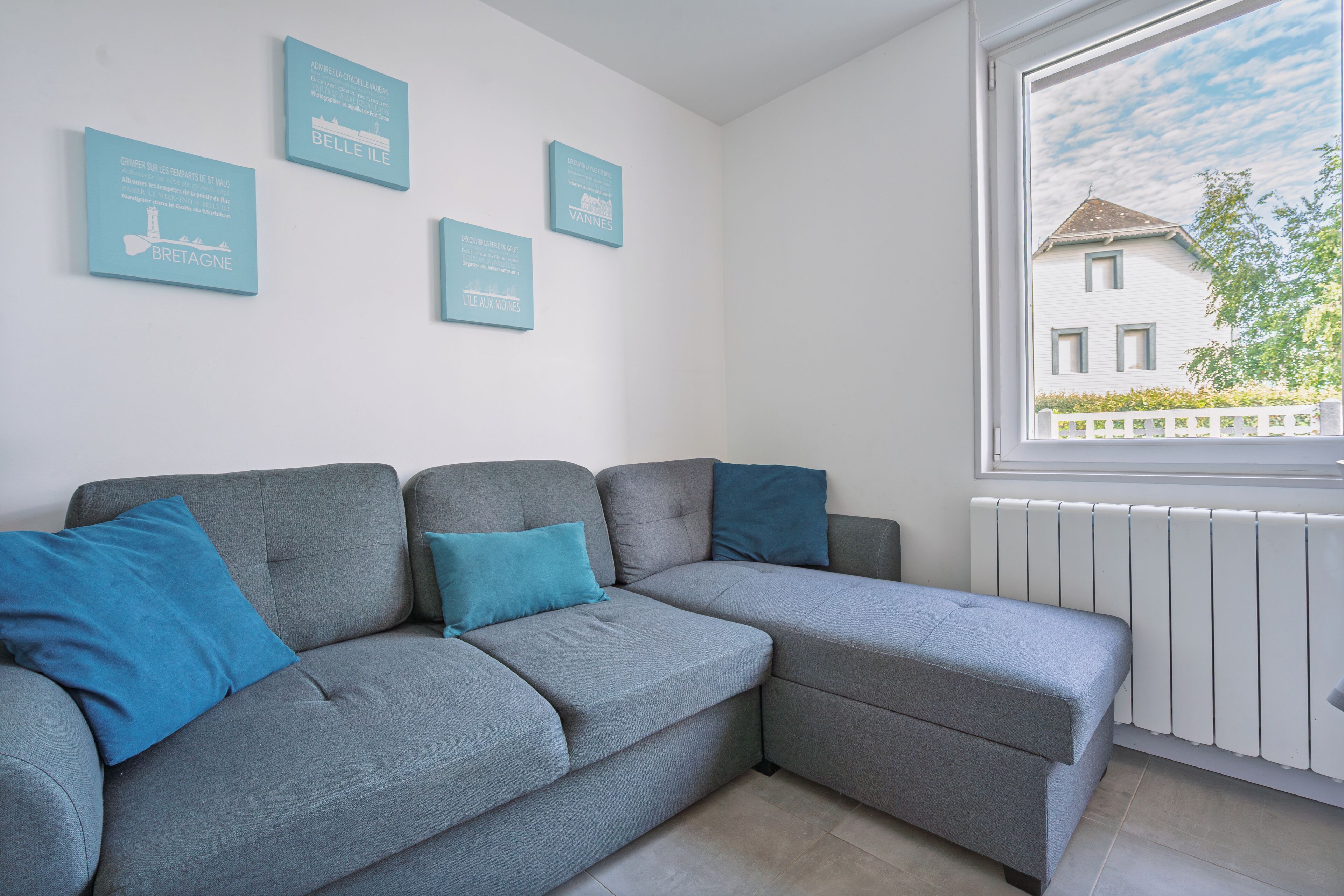 A grey sofa with blue cushions, a white wall with three framed signs, and a window with a view of a house.
