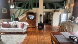 A spacious living room with a wooden floor, a white sofa, a dining table with chairs, a fireplace, and a staircase with green railings. - Charming 2-bedroom cottage w WiFi, AC in tranquil Syria w deck sounds Rose River (Syria)