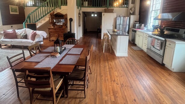A spacious kitchen with wooden flooring, a dining area with a table and chairs, a staircase with green railings, and a kitchen island with a refrigerator and stove. - Charming 2-bedroom cottage w WiFi, AC in tranquil Syria w deck sounds Rose River (Syria)