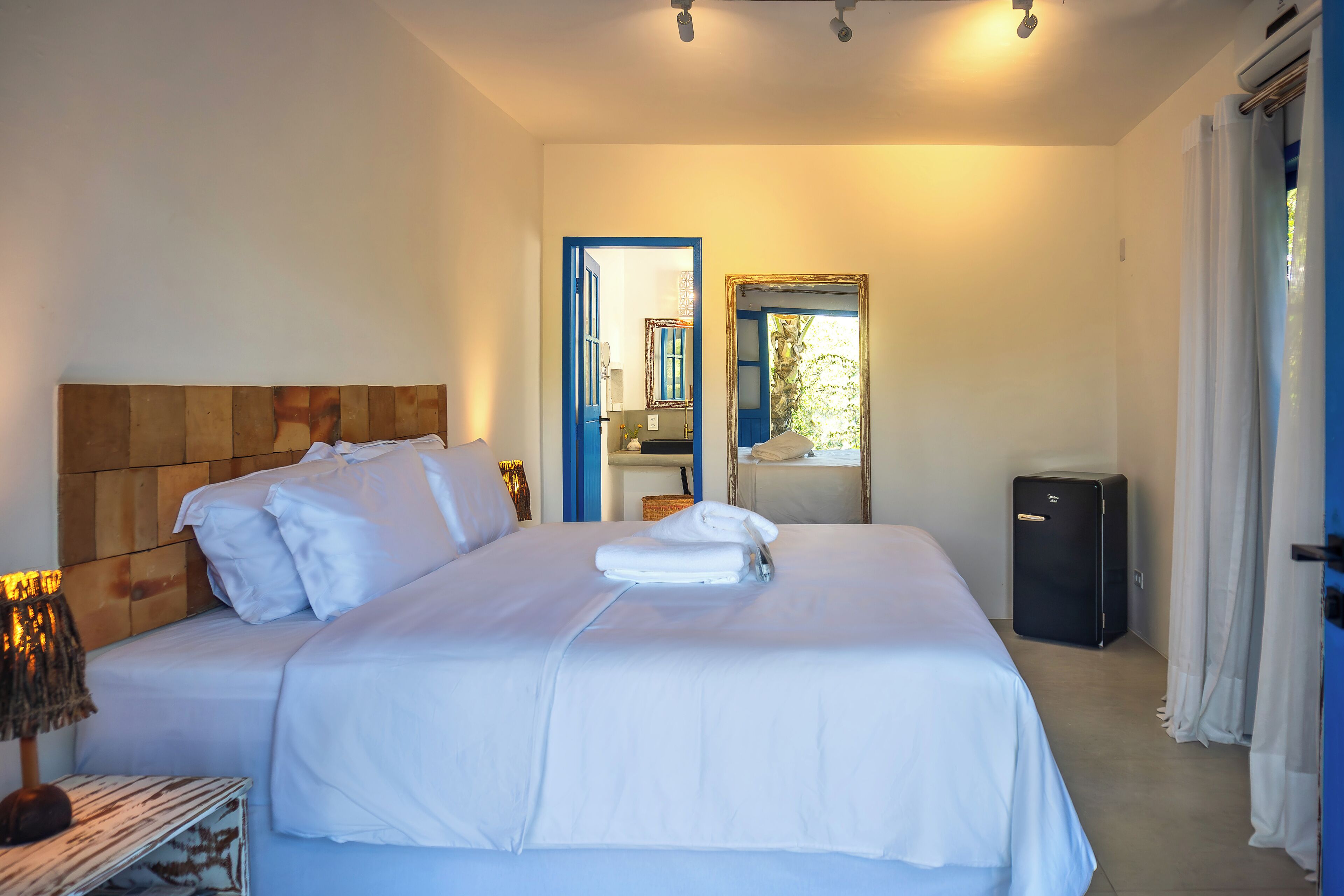 Una habitación de hotel con cama, mesita de noche con lámpara, espejo y maleta.