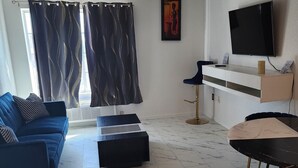A modern living room with a blue sofa, a black ottoman, a marble floor, a TV mounted on the wall, and a window with curtains. - The Enchanted Suites & Residence  (Roswell)