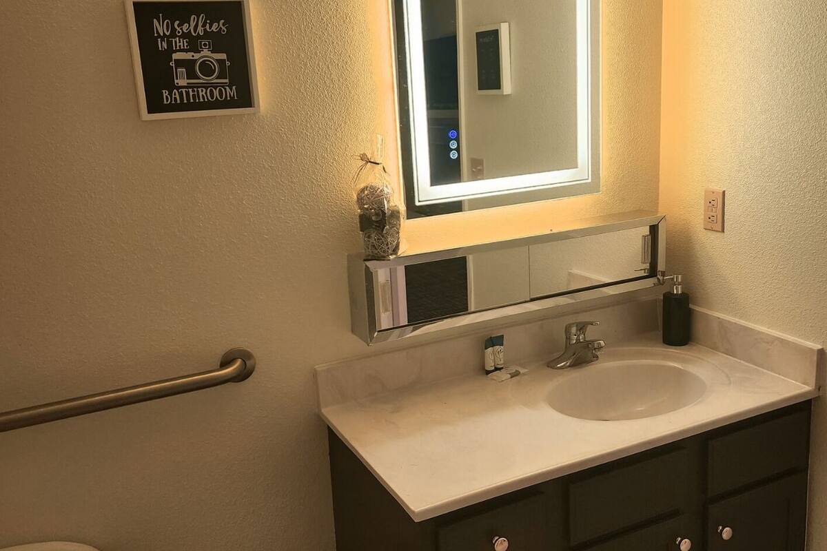 Uma casa de banho com um espelho grande, duas pias, um sanita e um cesto de lixo.