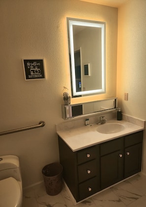 A bathroom with a large mirror, two sinks, a toilet, and a trash can. - The Enchanted Suites & Residence  (Roswell)