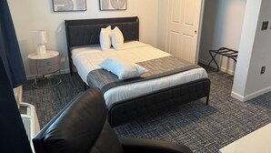 A hotel room with a bed, a nightstand, a chair, a wardrobe, and two abstract paintings on the wall. - The Enchanted Suites & Residence  (Roswell)